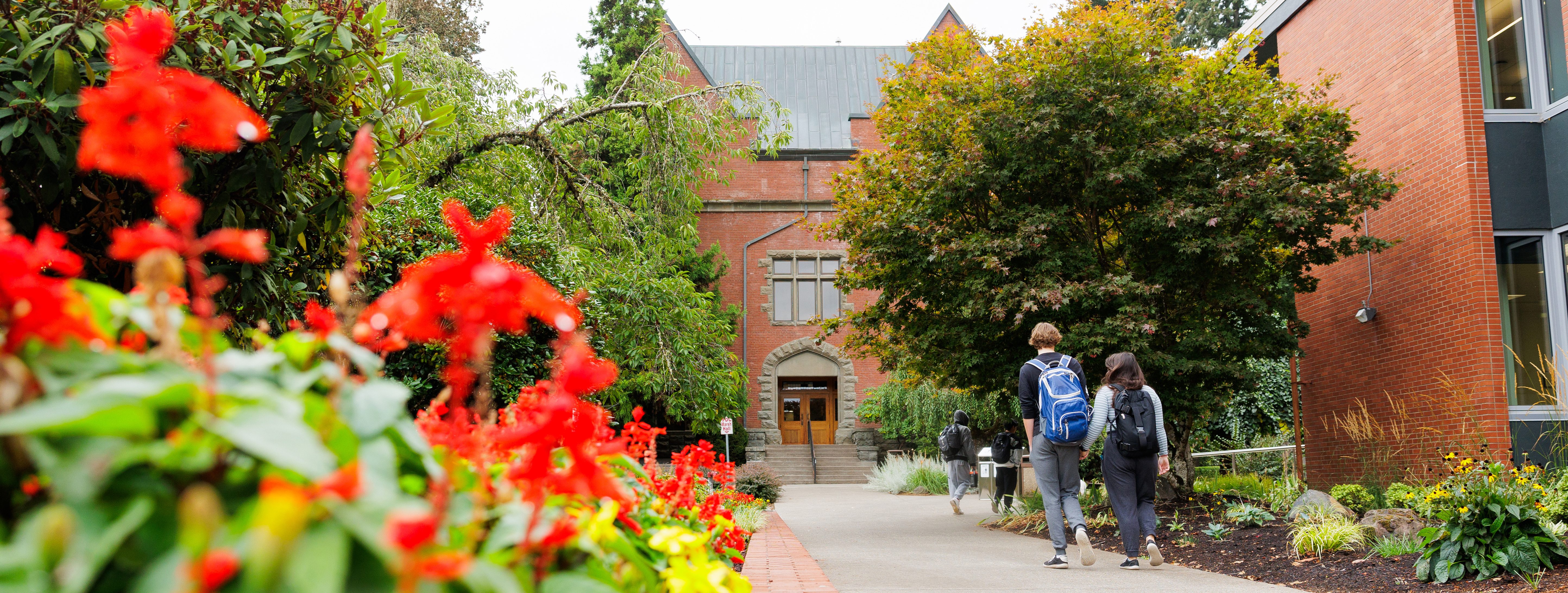 Students wearing backpacks walk toward Marsh Hall with red blooming flowers in the foreground.